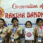 Four young girls in beige school uniforms with lanyards stand in front of a rakshabandhan banner proudly displaying handmade bracelets and crafts